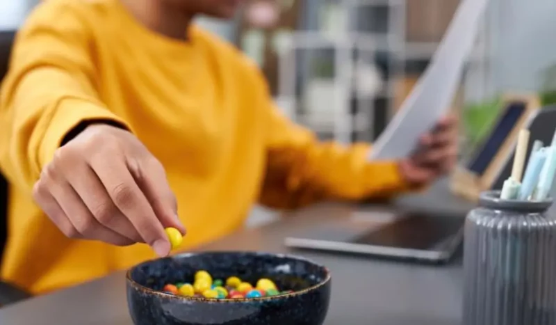 Person in yellow sweater grabbing colorful snacks from a bowl while reading a document at a desk, illustrating shifting consumer snacking habits and multitasking during work hours in 2025.