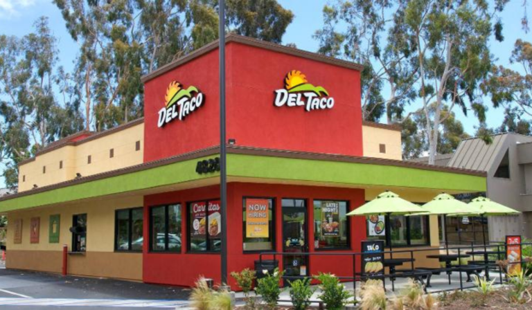 Street-facing view of a Del Taco restaurant with bright red and green exterior, outdoor seating, and branded signage.