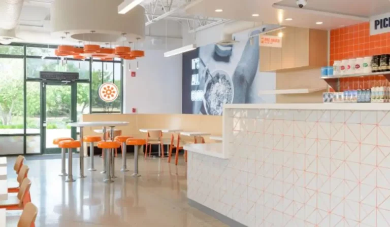 Interior view of a Protein Bar & Kitchen fast-casual restaurant, featuring a clean, modern dining space with orange seating, white countertops, and branded pickup area.