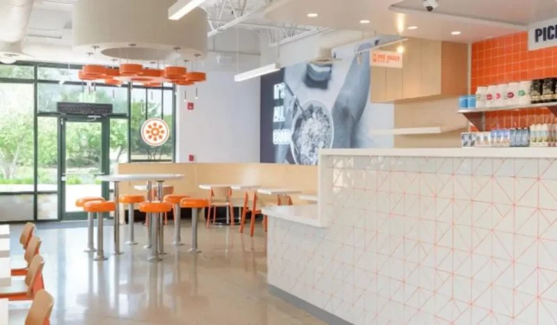 Interior view of a Protein Bar & Kitchen fast-casual restaurant, featuring a clean, modern dining space with orange seating, white countertops, and branded pickup area.